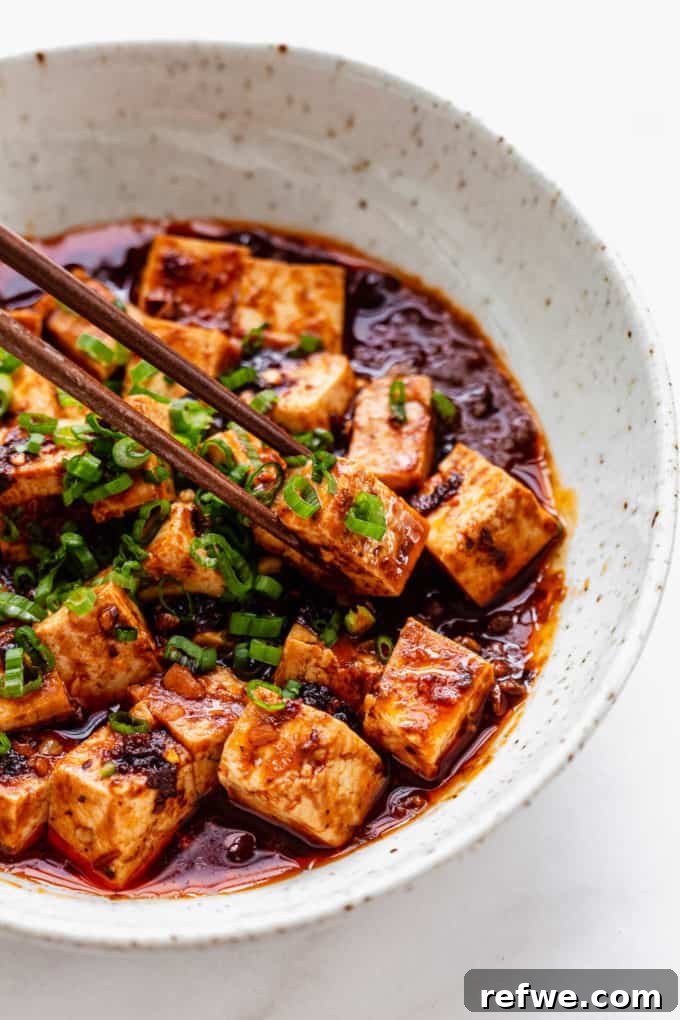 Close-up of chopsticks expertly picking up a succulent cube of Vegetarian Mapo Tofu, perfectly coated in the rich, spicy, and glossy sauce.