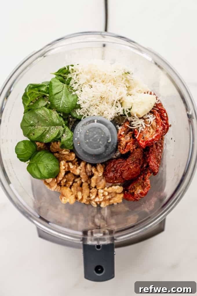 Velvety Red Pesto Pasta 3 Sun-dried tomatoes, parmesan cheese, walnuts, and basil leaves being added into a food processor for pesto preparation.