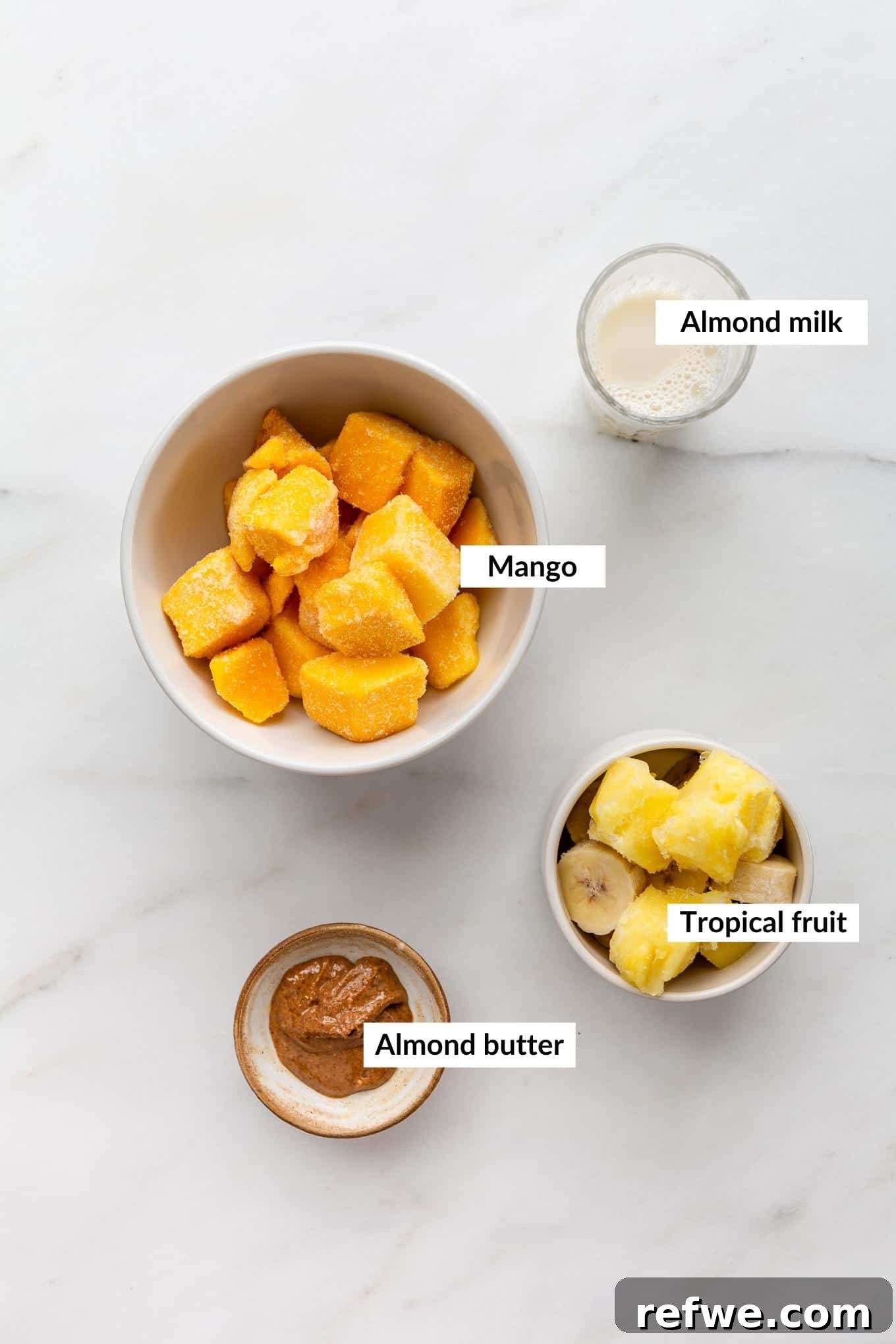 Golden Mango Bliss Bowl 3 A flat lay image displaying the key ingredients for a mango smoothie bowl: a pile of vibrant frozen mango chunks, slices of frozen banana, a glass of almond milk, and a spoonful of creamy almond butter, arranged on a light surface.