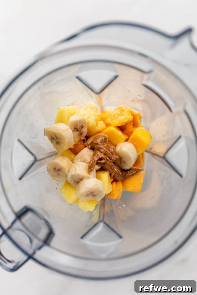 Golden Mango Bliss Bowl 4 Close-up of frozen mango, banana, pineapple chunks, and a dollop of almond butter being added into a high-speed blender, ready to be transformed into a creamy smoothie bowl.