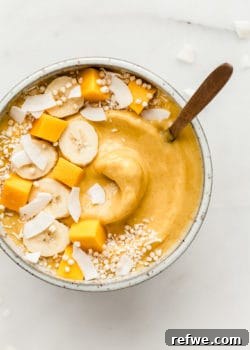 A mango smoothie bowl artfully topped with fresh fruits like sliced mango, banana, kiwi, and passionfruit, alongside crunchy granola and delicate coconut flakes.
