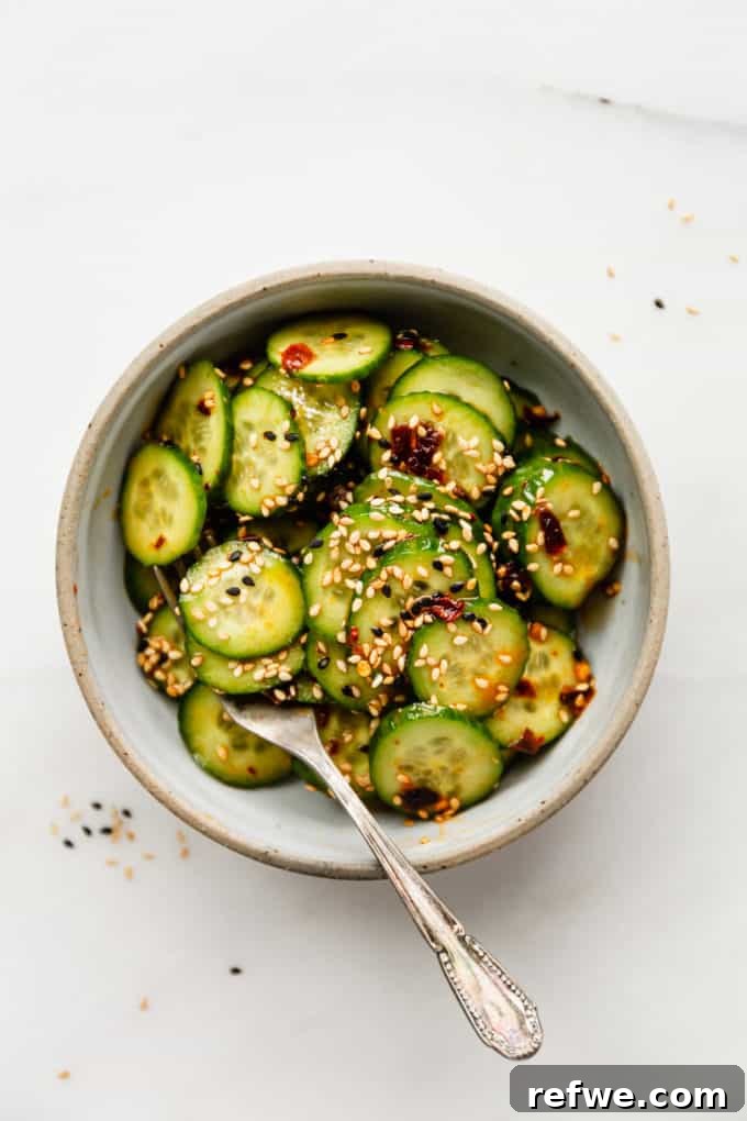 A close-up shot of thinly sliced cucumbers arranged in a sleek white bowl, drizzled with a rich amber Asian dressing and garnished with toasted sesame seeds, highlighting its fresh appeal.