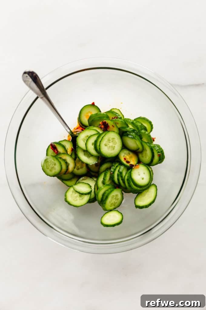 Thinly sliced cucumbers resting at the bottom of a mixing bowl, awaiting a generous pour of the homemade Asian dressing, ready to be tossed.