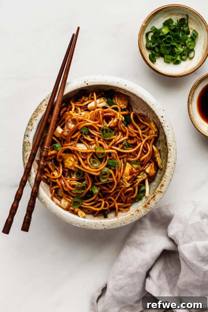 Indonesian Fried Noodles Mie Goreng 15 A beautifully plated bowl of Mie Goreng noodles, ready to eat, with a grey napkin and a small bowl of green onions on the side.