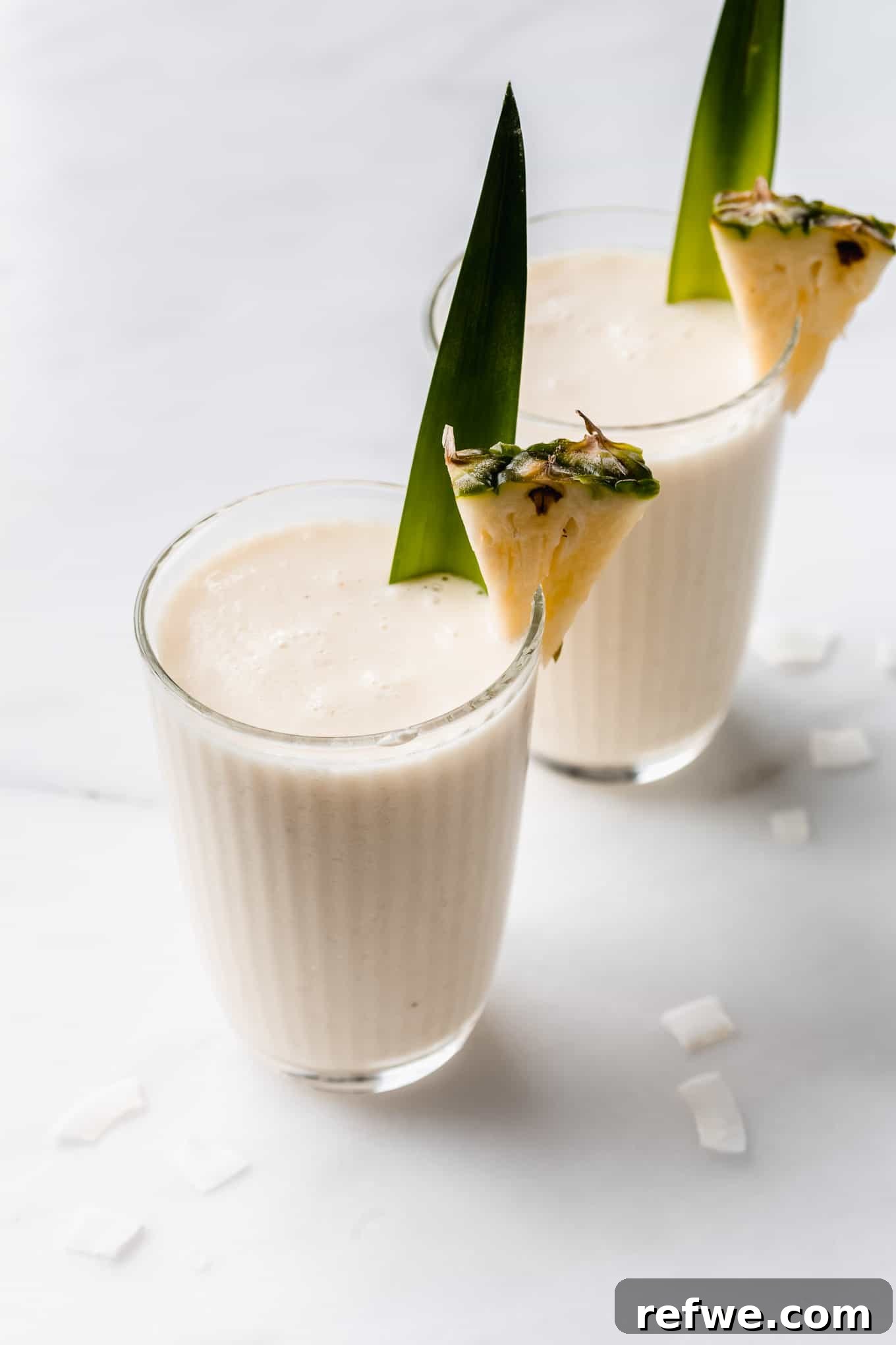 Pina Colada Dream Smoothie 2 Two glasses of refreshing Pina Colada Smoothies, garnished with pineapple slices and tropical leaves, set against a bright, summery background.