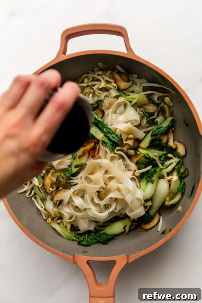 Wok-Tossed Ho Fun 8 Rich stir-fry sauce being poured over a harmonious blend of noodles and vegetables in a pan, about to be mixed.