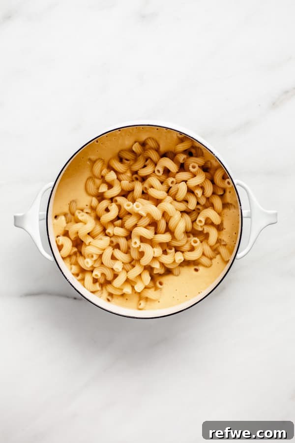 A white pot containing the creamy cheese sauce, with al dente macaroni noodles recently added, awaiting a thorough mix.