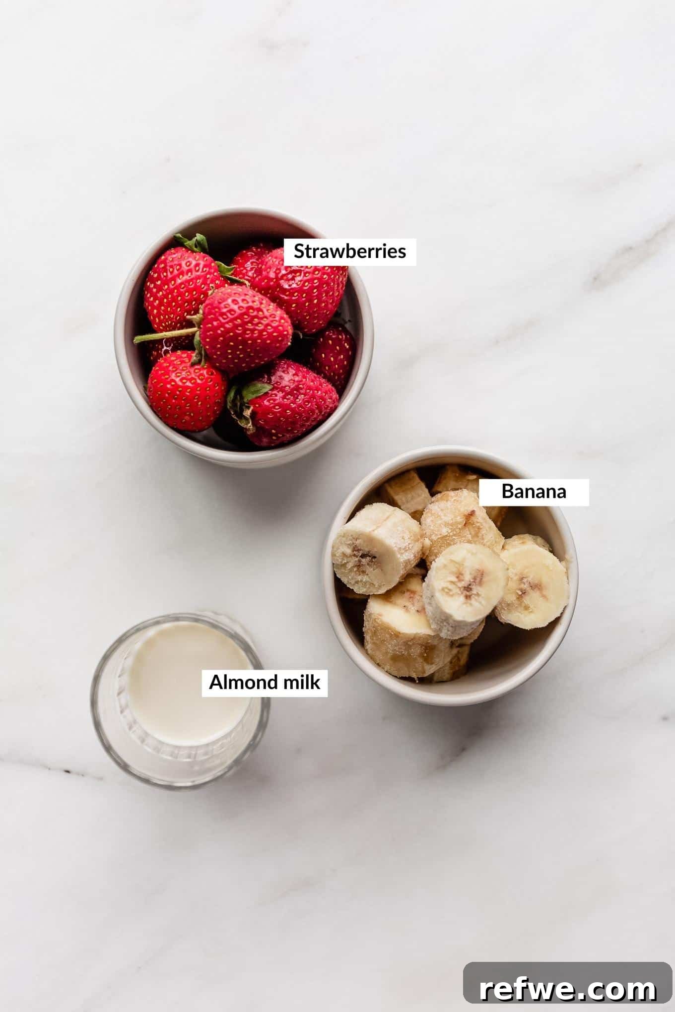 Strawberry Bliss Bowl 3 A flat lay image displaying the simple ingredients required for a strawberry smoothie bowl: fresh strawberries, sliced frozen bananas, and a carton of almond milk.