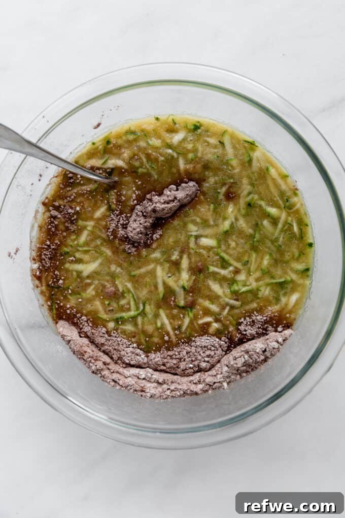 Wholesome Cocoa Zucchini Muffins 7 Wet ingredients with zucchini being gently mixed into the dry flour mixture in a large mixing bowl.