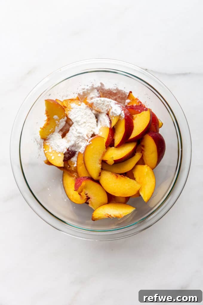 Sliced peaches being mixed with cornstarch in a clear bowl, preparing for the crisp.