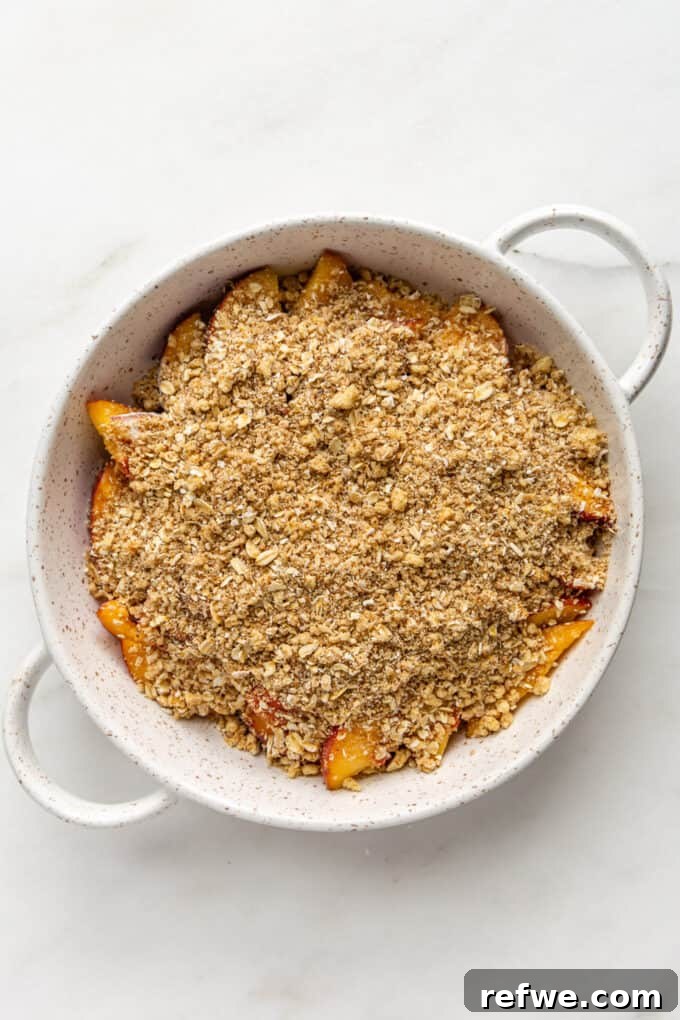 The crumbly crisp topping evenly sprinkled over the peaches in a white baking dish, ready for baking.