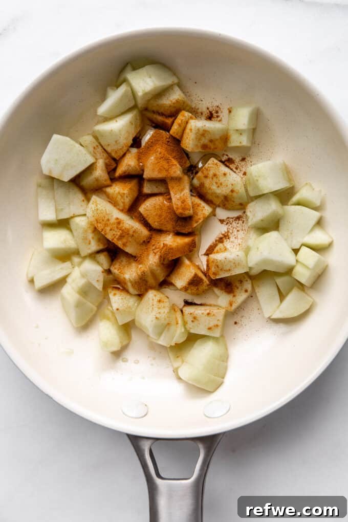 No-Bake Apple Pie Oats 8 Peeled and cubed apples mixed with cinnamon and maple syrup in a white non-stick pan, ready for cooking.
