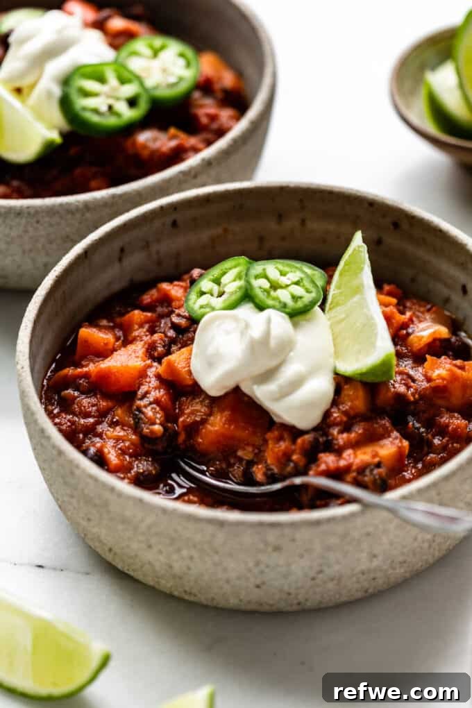 Hearty Sweet Potato Black Bean Chili 12 A close-up shot of a steaming bowl of sweet potato chili, topped generously with creamy sour cream, vibrant jalapeño slices, and a fresh lime wedge, resting on a rustic wooden surface.
