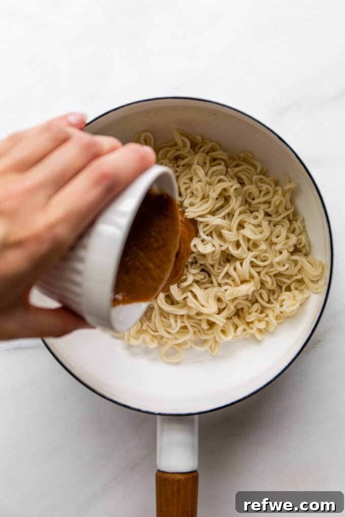 A hand pouring rich, freshly made peanut butter sauce into a pot of cooked ramen noodles.