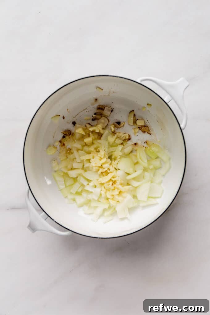 Chopped onions and minced garlic gently sautéing in avocado oil within a white pot, forming the aromatic base of the chili.