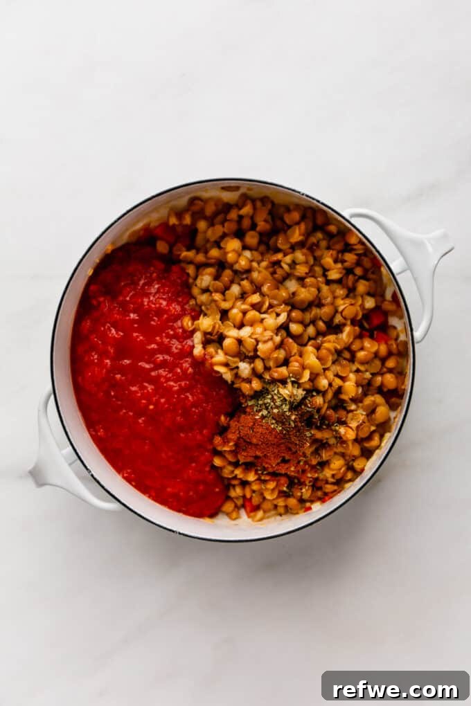 Drained lentils, rich crushed tomatoes, and a colorful array of spices added to the sautéed vegetables in a white pot, ready for simmering.
