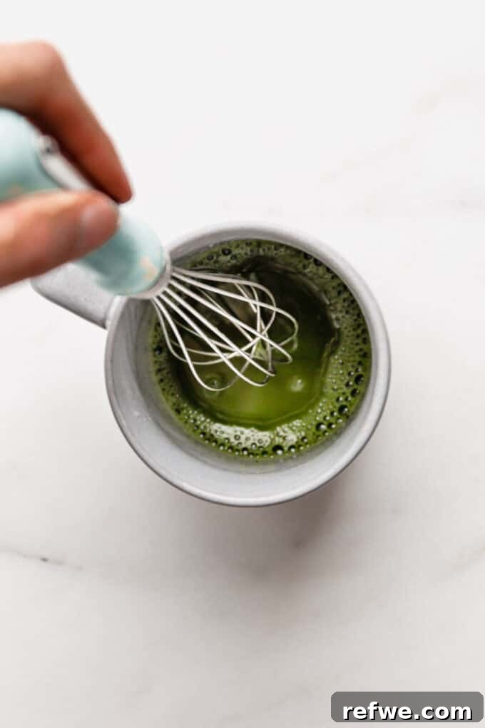 A whisk vigorously blending matcha powder and hot water in a mug, creating a smooth green paste.