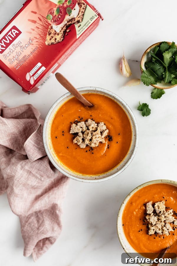 Hearty Spiced Sweet Potato, Lentil, and Carrot Soup 3 A beautifully styled bowl of sweet potato and lentil soup on a marble board, accompanied by a pink napkin, a small bowl of cilantro, and a box of Ryvita crispbreads, highlighting its fresh ingredients and perfect pairing.