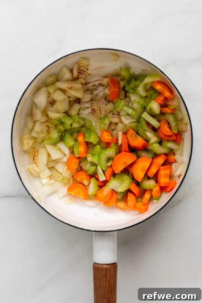 A mix of carrots, celery, and onion sautéing in a white pot, forming the colorful and aromatic base of the soup.