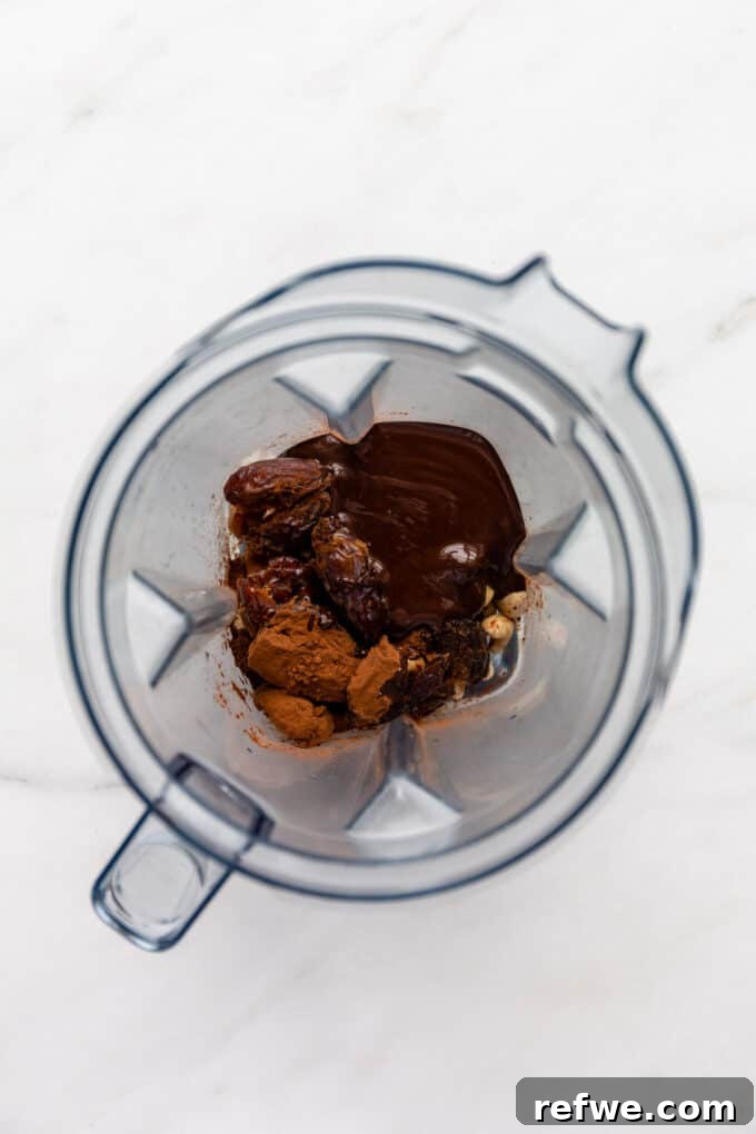 All dry and liquid ingredients, including cashews, dates, cocoa powder, and melted dark chocolate, gathered in the blender jar, prepared for the next step.