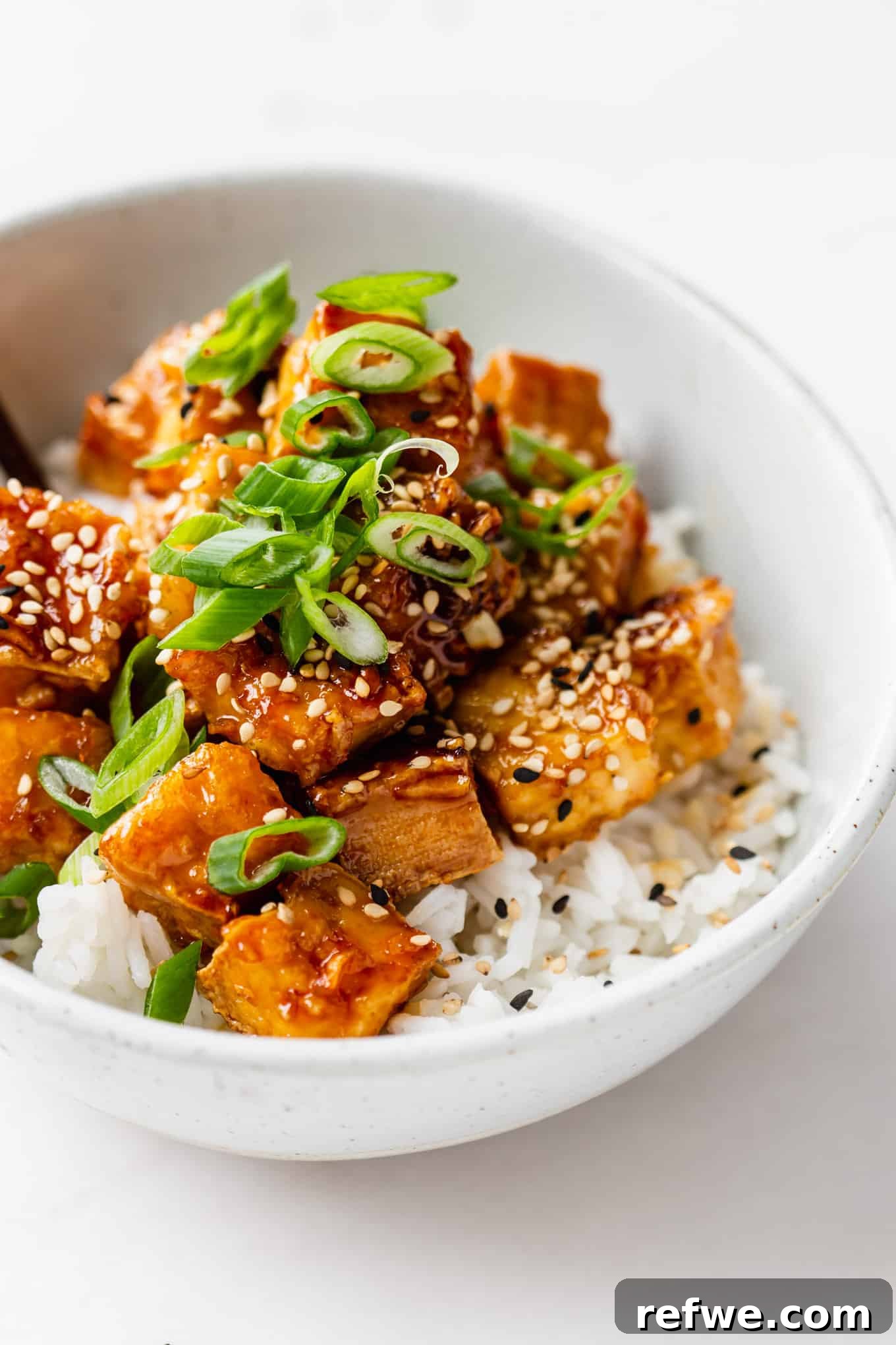 A vibrant bowl of crispy honey garlic tofu served over fluffy white rice, garnished with fresh green scallions and a sprinkle of toasted sesame seeds.