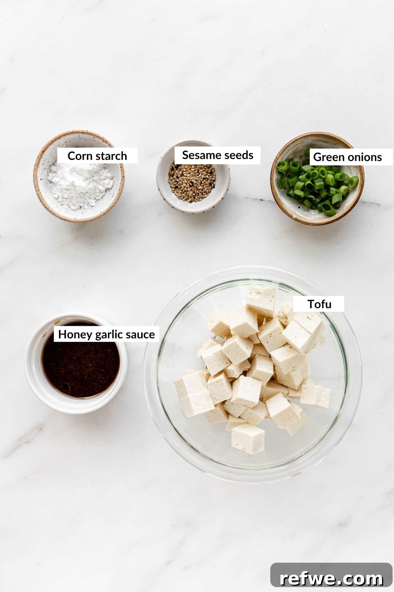 A collection of fresh ingredients laid out, including a block of tofu, a small bowl of cornstarch, fresh ginger, garlic cloves, honey, soy sauce, and a bottle of sriracha, all ready for making honey garlic tofu.