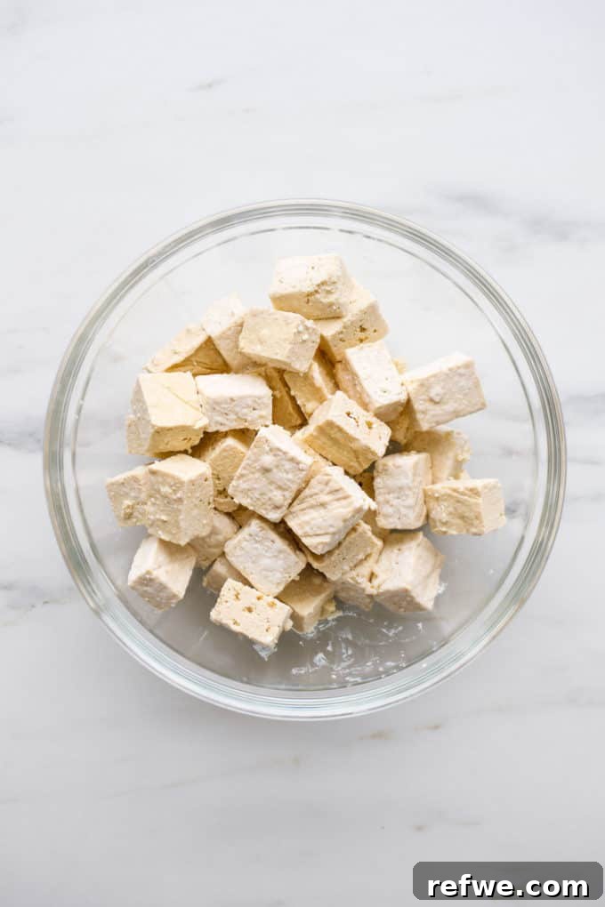 Cubed tofu, shimmering slightly with oil, being gently tossed in a clear mixing bowl to coat each piece thoroughly with fine white cornstarch.