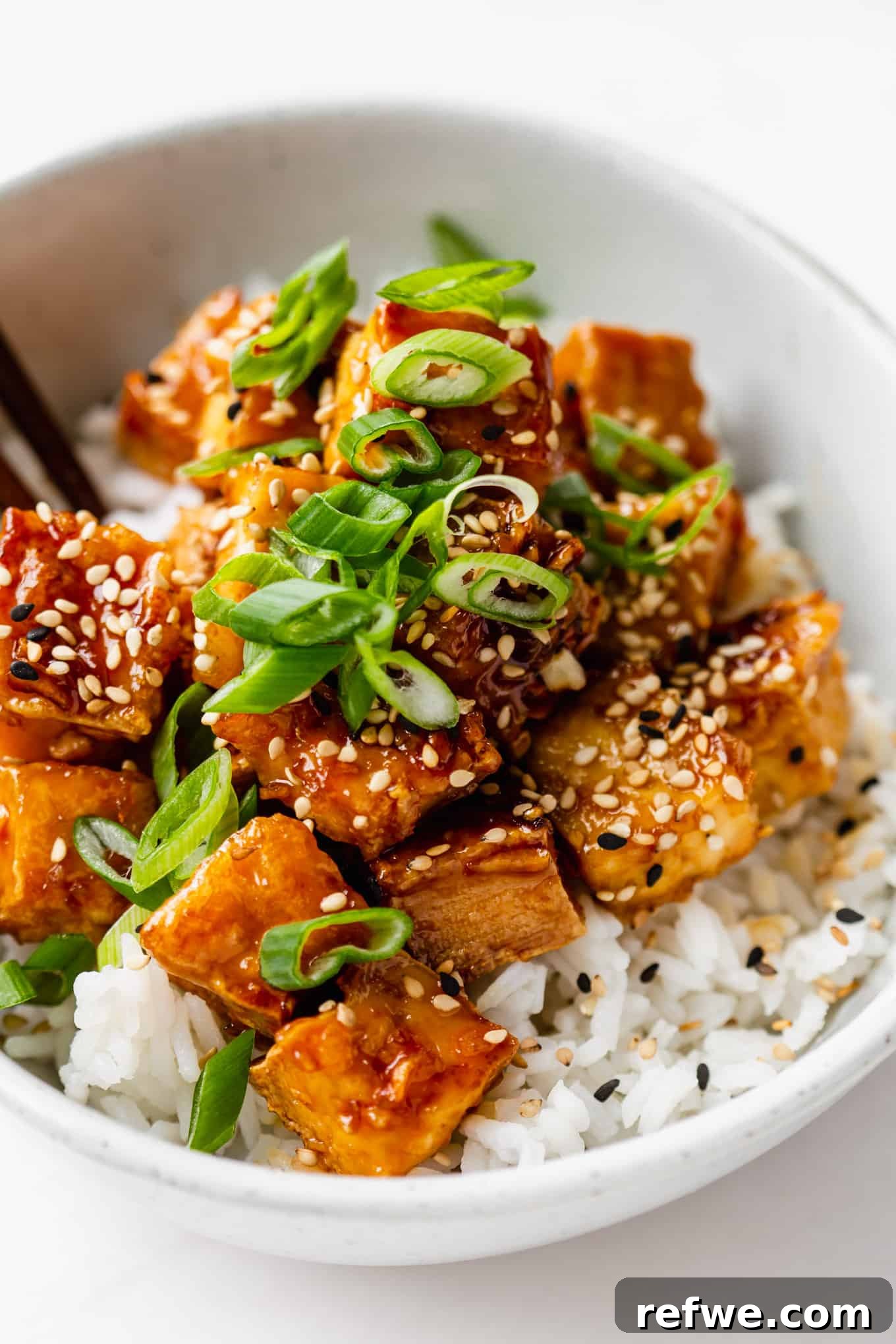 A close-up shot of a serving of honey garlic tofu generously topped with vibrant green scallions and a scattering of white sesame seeds, showcasing its appealing texture and glaze.