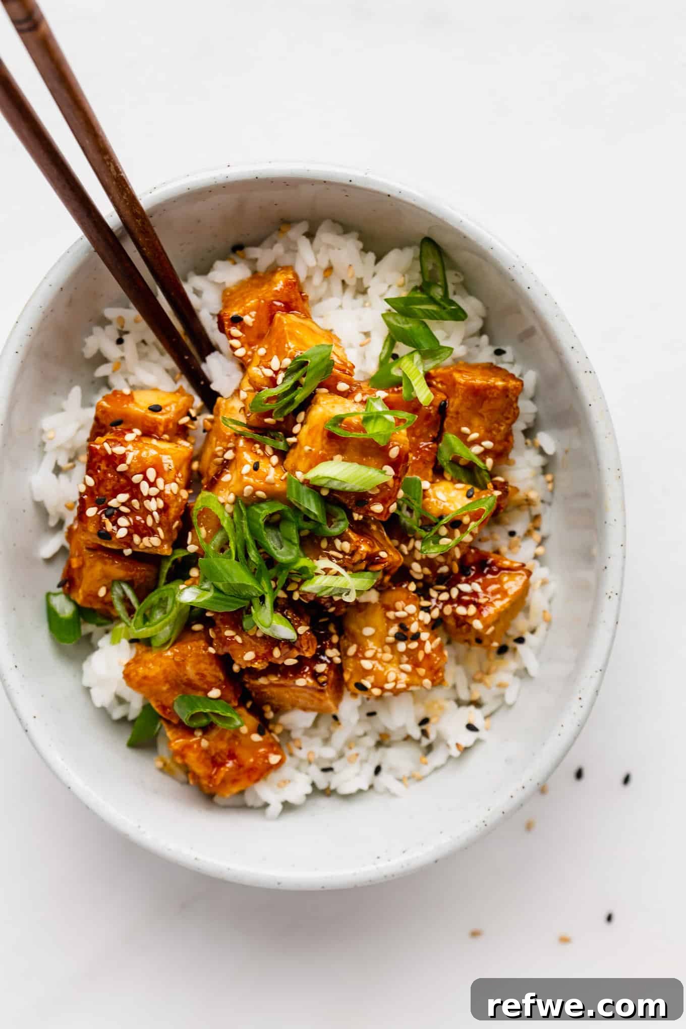 A beautifully plated bowl featuring honey garlic tofu and rice, perfectly set for enjoyment with a pair of elegant wooden chopsticks resting on the side.
