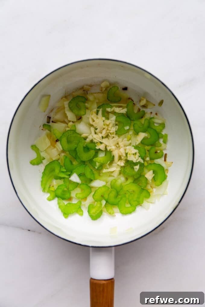 Sautéing Aromatics for Soup Base Finely chopped celery, garlic, and onion gently sautéing in oil within a white pot, forming the aromatic base of the soup.