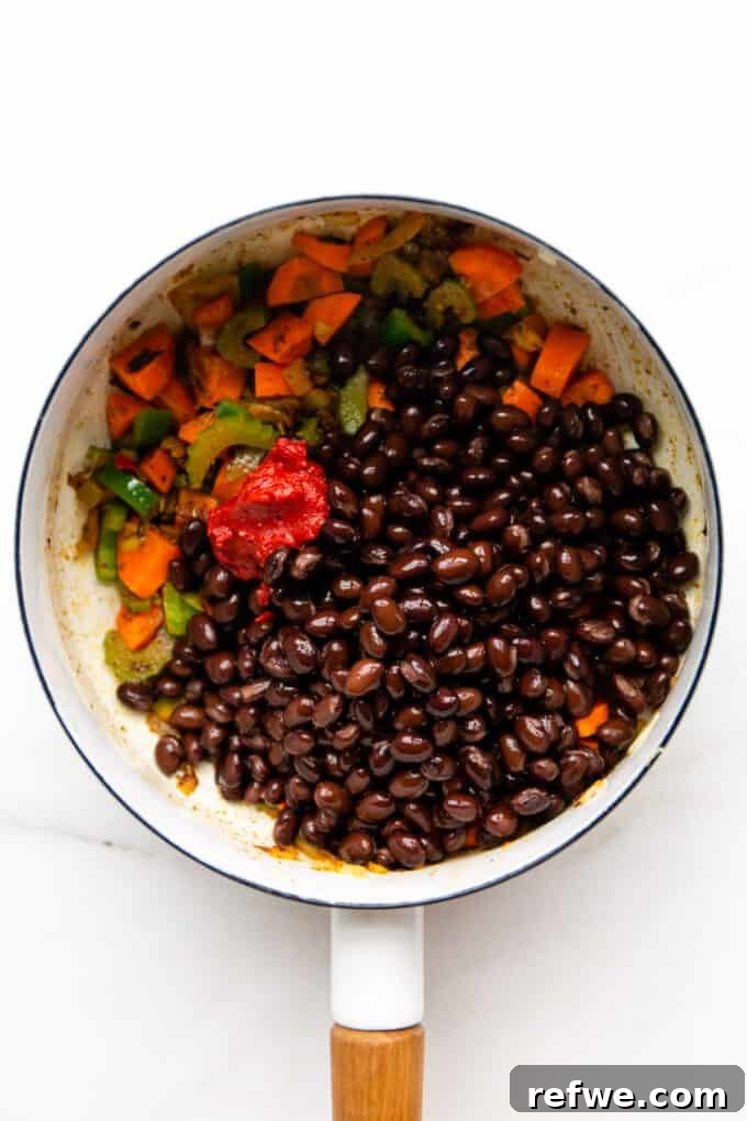 Combining Ingredients in Pot Black beans, sautéed vegetables, and tomato paste combined in a white pot, ready for the broth to be added.