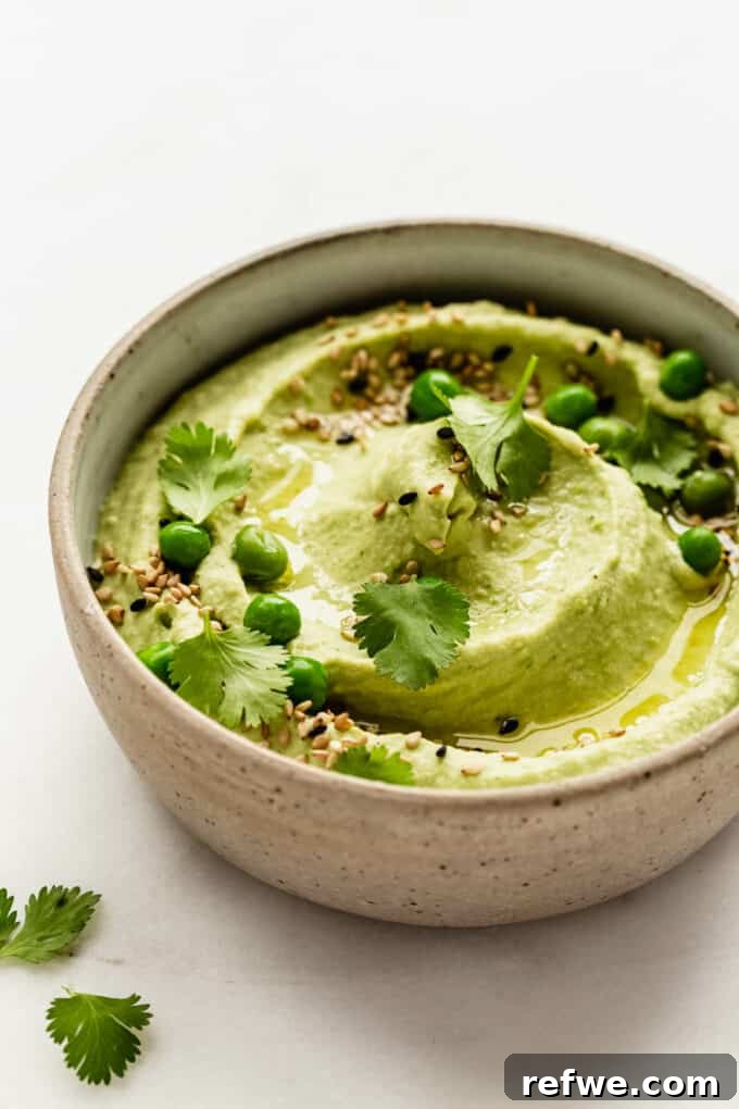 A ceramic bowl filled with vibrant green pea hummus, garnished with fresh cilantro and a few whole green peas.