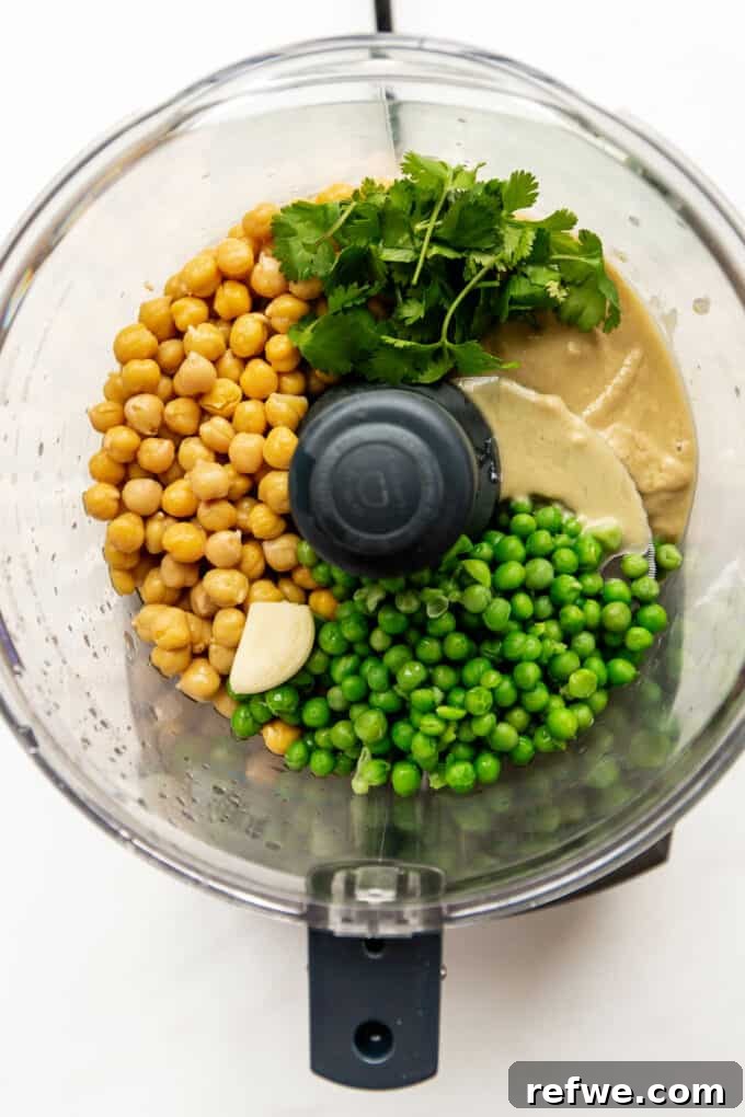 A food processor bowl containing drained chickpeas, blanched green peas, fresh cilantro, and whole garlic cloves, ready for blending.