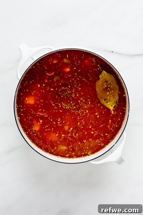 A white pot containing vegetable broth and rich crushed tomatoes, creating the foundation of the minestrone soup.