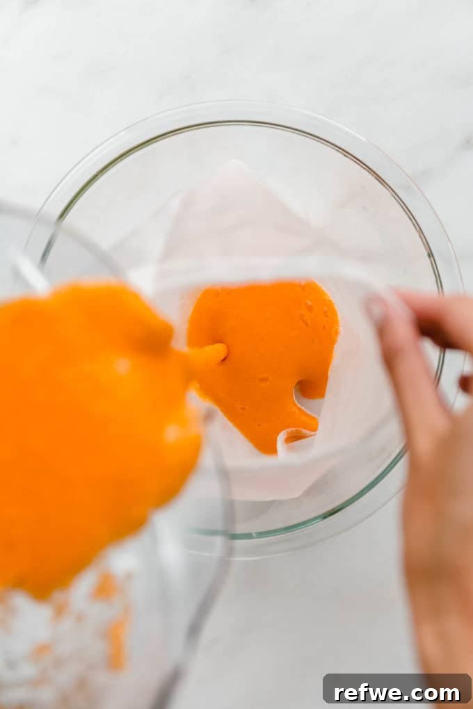 Zingy Ginger Carrot Orange Immunity Nectar 6 A hand pouring the blended orange, carrot, and ginger mixture into a mesh straining bag held over a mixing bowl, extracting the pure juice.