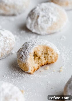 A close-up of an almond snowball cookie with a small bite taken out, showing the tender crumb and snowy powdered sugar coating.