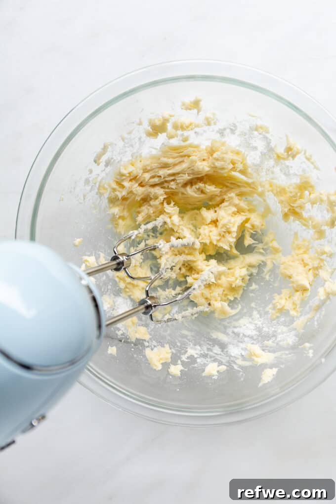 A hand mixer blending softened butter and powdered sugar in a bowl, creating a light and fluffy consistency.
