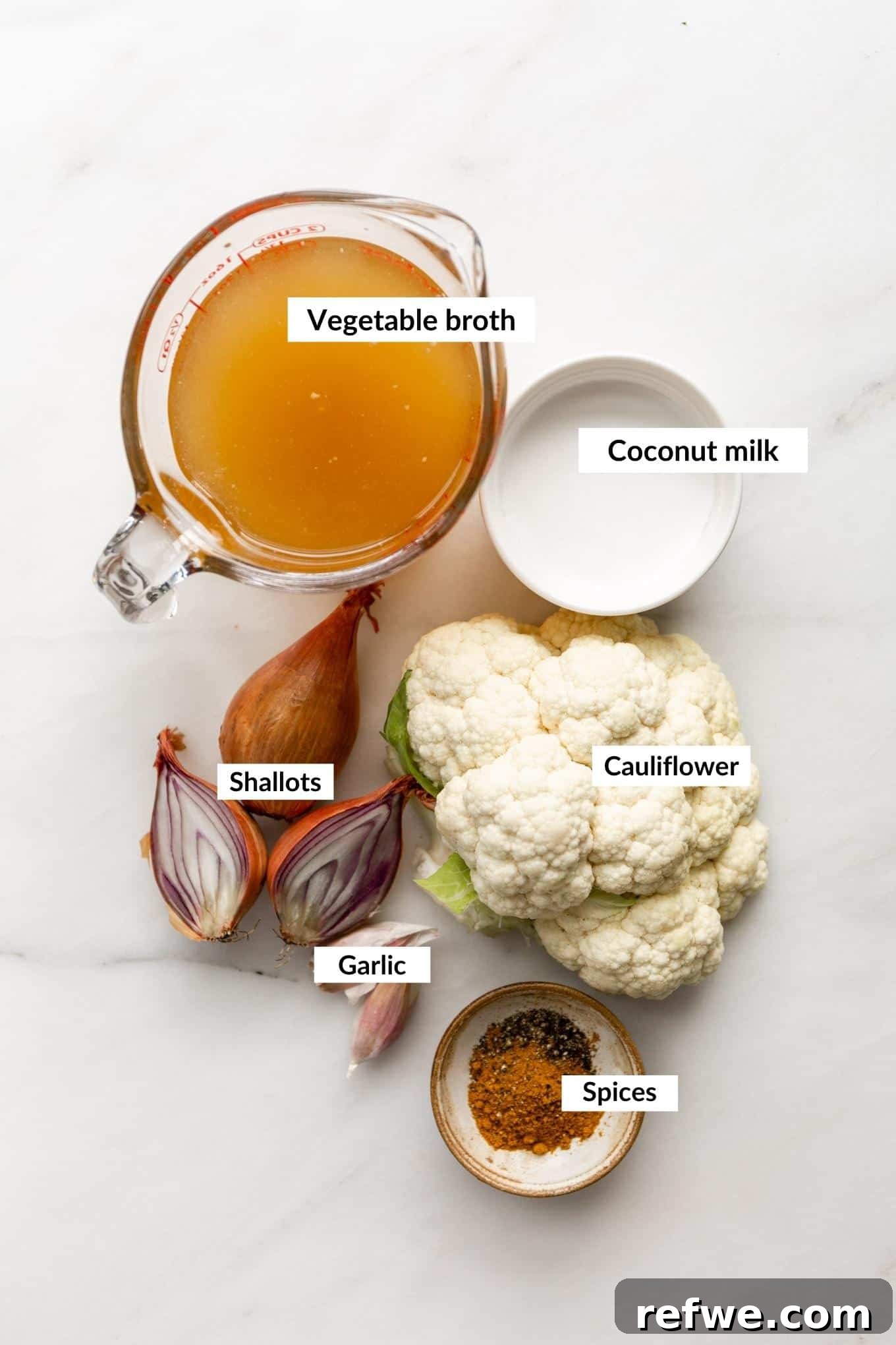 Velvety Curried Cauliflower Bliss 3 A flat lay photograph showcasing the key ingredients for curried cauliflower soup: cauliflower florets, shallots, garlic, a can of coconut milk, and a jar of curry powder.