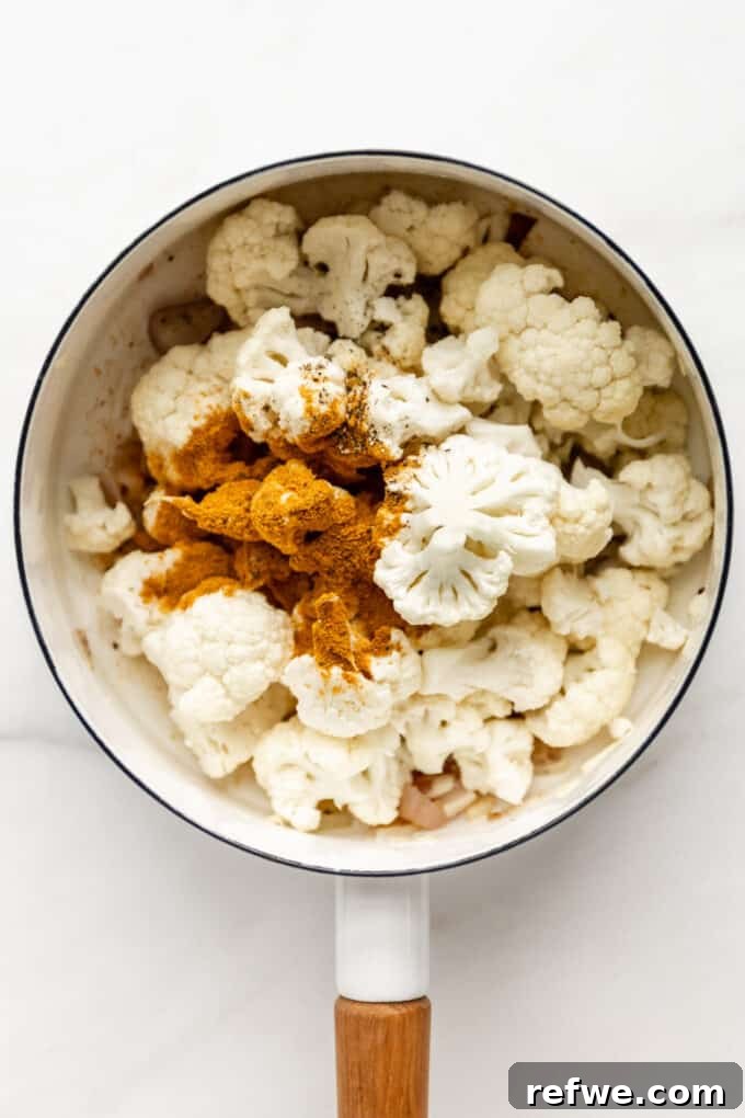 Velvety Curried Cauliflower Bliss 5 Cauliflower florets and curry powder being stirred into the sautéed aromatics in a white pot.