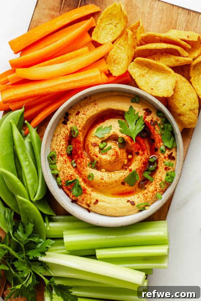 Creamy Buffalo hummus served on a rustic wood board, surrounded by fresh vegetables like celery and carrots, and crispy pita chips, highlighting its vibrant color and tempting texture.