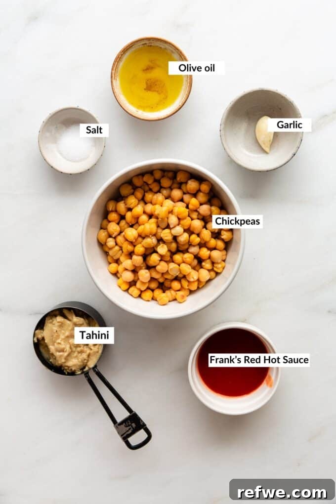 A vibrant flat lay showcasing all the fresh ingredients for homemade buffalo hummus, including a can of chickpeas, a bottle of Frank's Red Hot Sauce, tahini, olive oil, garlic, and spices, neatly arranged on a white background.