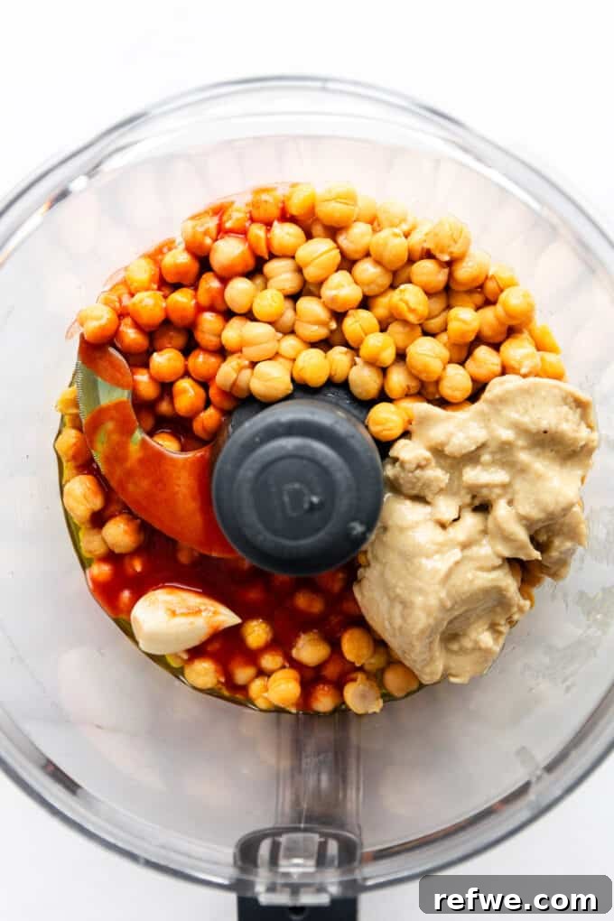 Close-up shot inside a food processor bowl, showing chickpeas, a rich red swirl of Frank's Red Hot Sauce, creamy tahini, and garlic cloves, ready to be blended into buffalo hummus.