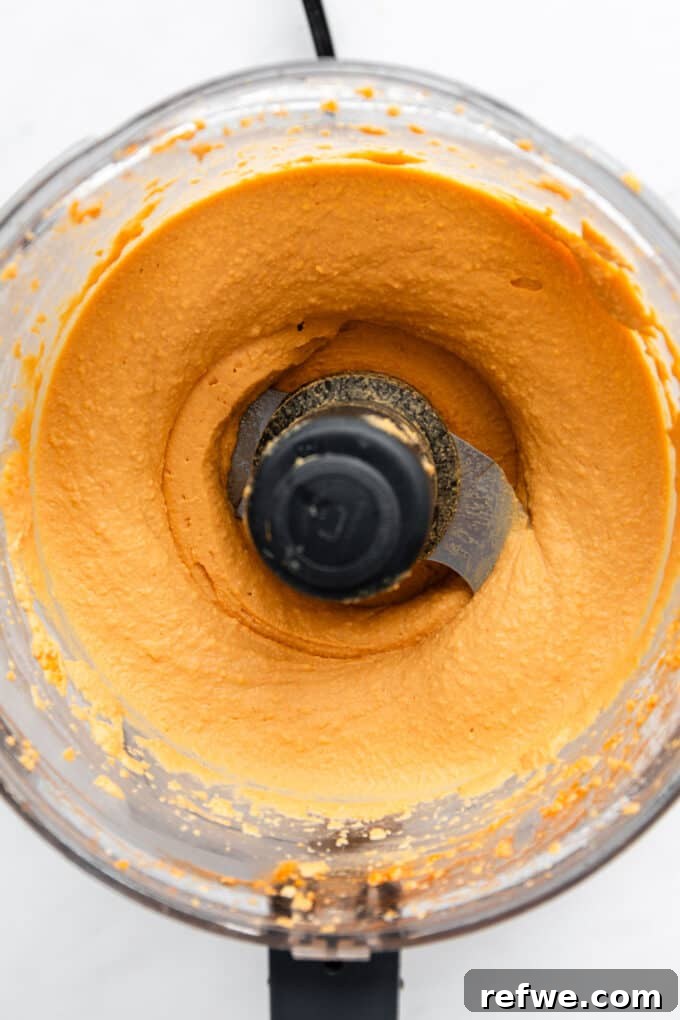 The finished buffalo hummus, perfectly blended to a smooth, creamy texture, fills the bowl of a food processor, ready to be transferred for serving.