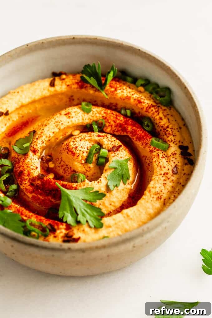 A beautifully presented bowl of buffalo hummus, generously topped with a sprinkle of smoked paprika, vibrant green onion, and fresh parsley, showcased on a textured surface.