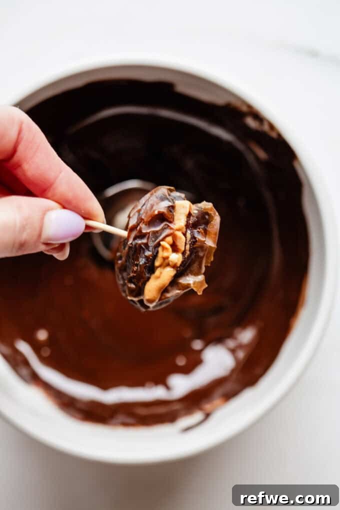 Snickers Dream Dates 7 A Medjool date, expertly stuffed with peanut butter and peanuts, being carefully dipped into a bowl of rich, melted dark chocolate, capturing the moment of transformation into a Date Snickers.