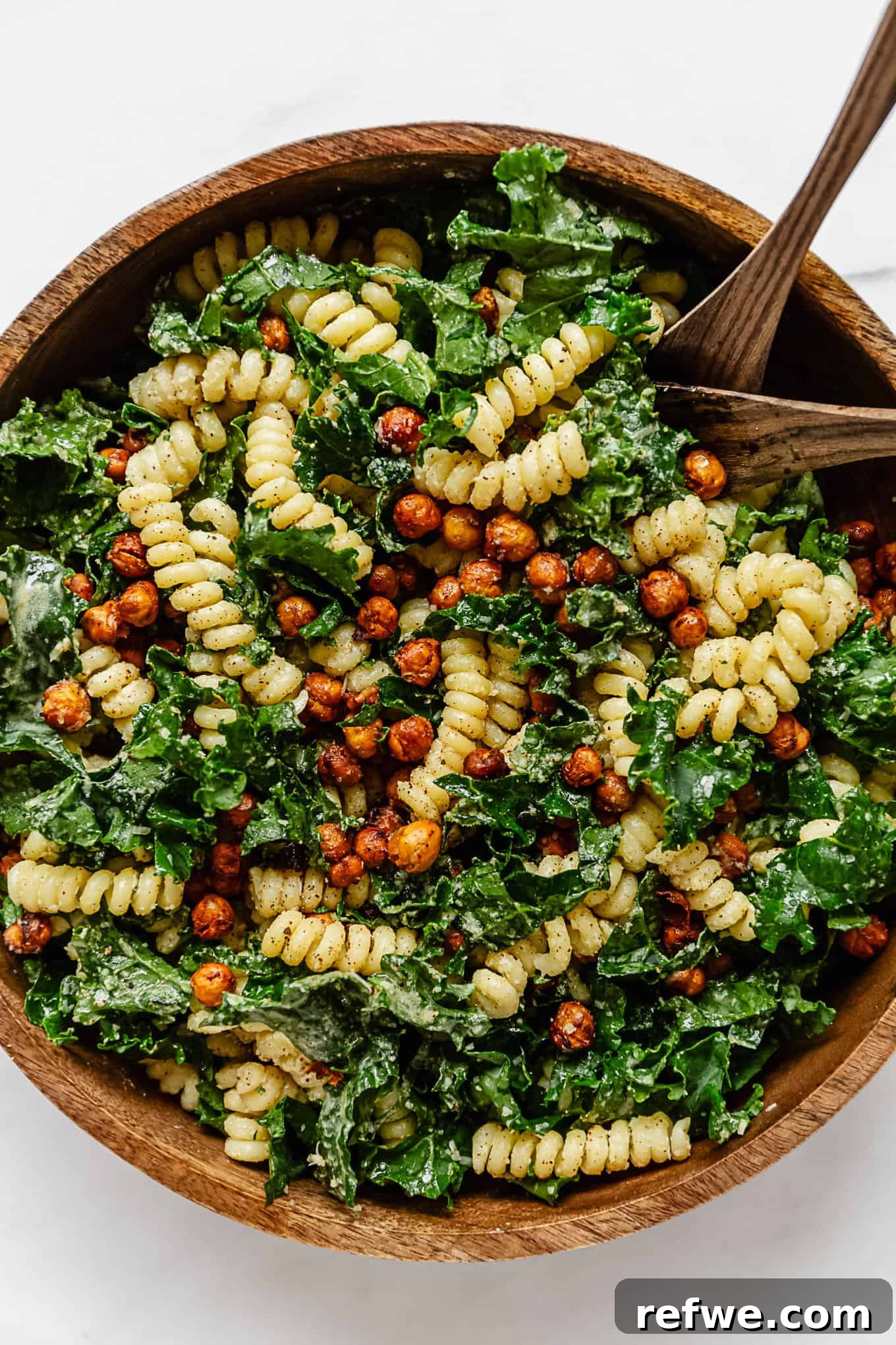 Caesar's Hearty Kale Pasta 12 A large wooden mixing bowl brimming with Kale Caesar Pasta Salad, accompanied by two wooden serving spoons.