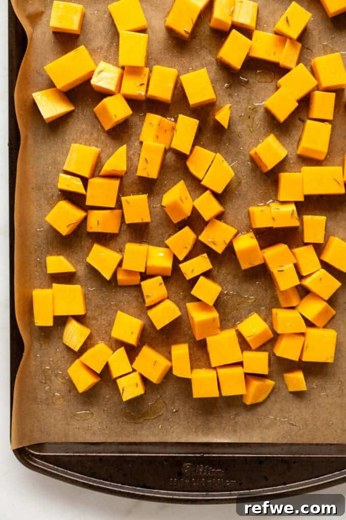 Cubed butternut squash seasoned with olive oil, thyme, salt, and pepper, spread evenly on a baking sheet, ready for roasting.