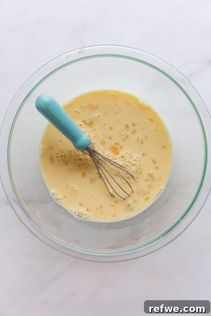 The liquid ingredients, eggs and milk, are vigorously whisked together in a mixing bowl using a blue whisk until thoroughly combined and smooth.