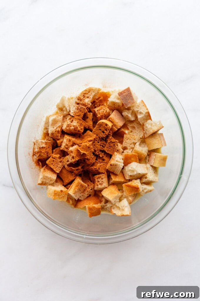Cubed bread pieces are mixed with a generous sprinkle of cinnamon in a mixing bowl, preparing them for soaking in the custard.