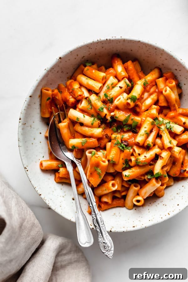 Velvety Vegan Roasted Red Pepper Pasta 5 Close-up of creamy vegan roasted red pepper pasta in a white bowl, with a fork and spoon, ready to be enjoyed.